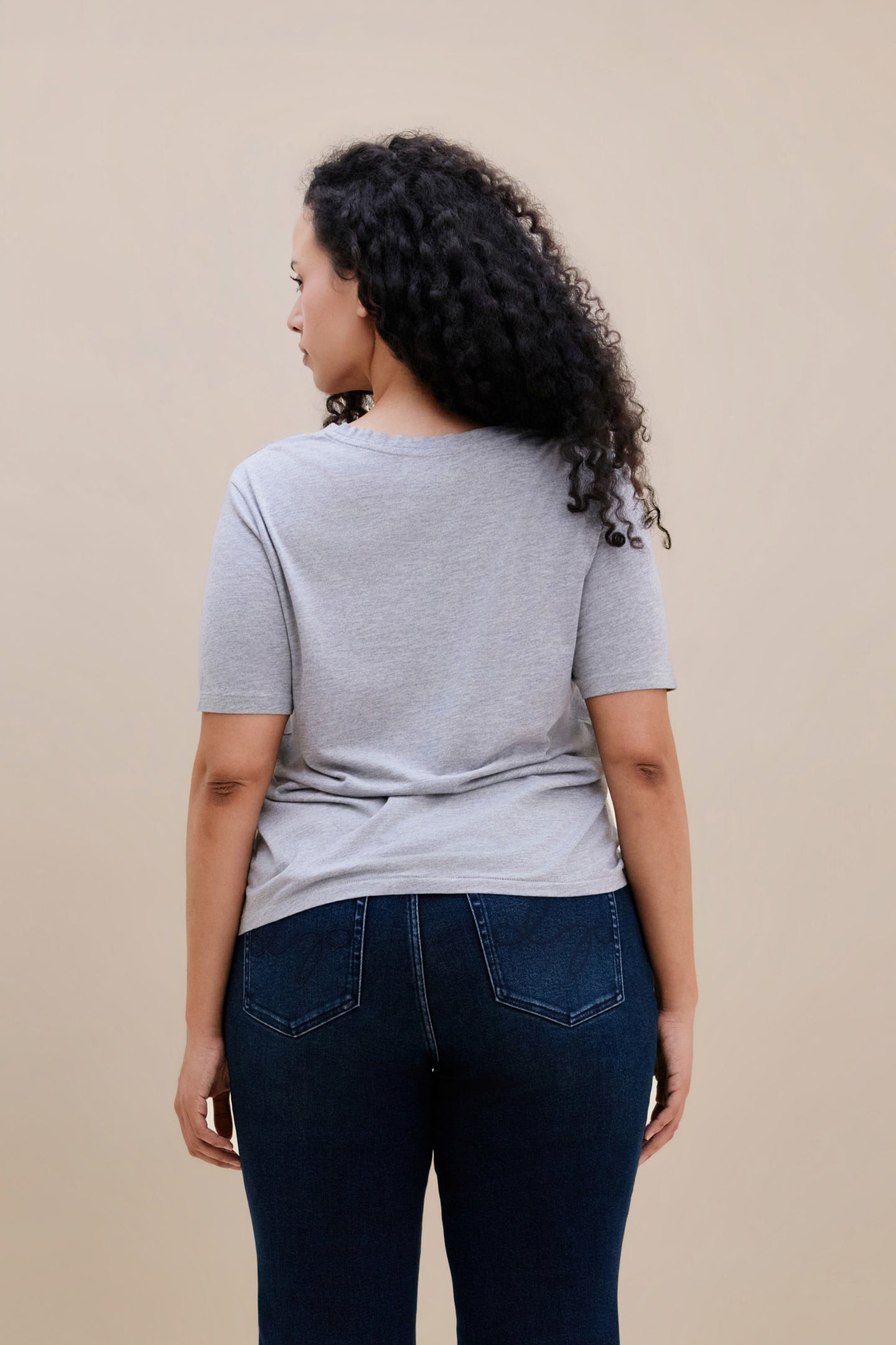 Backside of a woman wearing a heather grey v-neck t-shirt and dark jeans