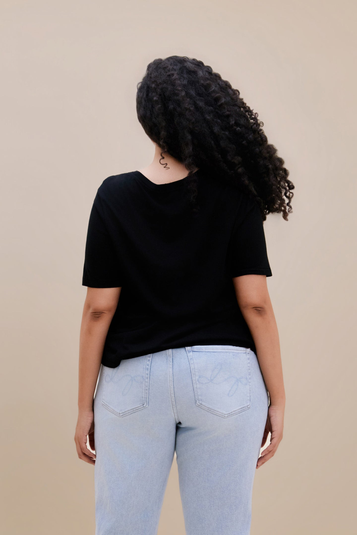 Backside of a woman wearing a black t-shirt and light blue jeans against a beige background