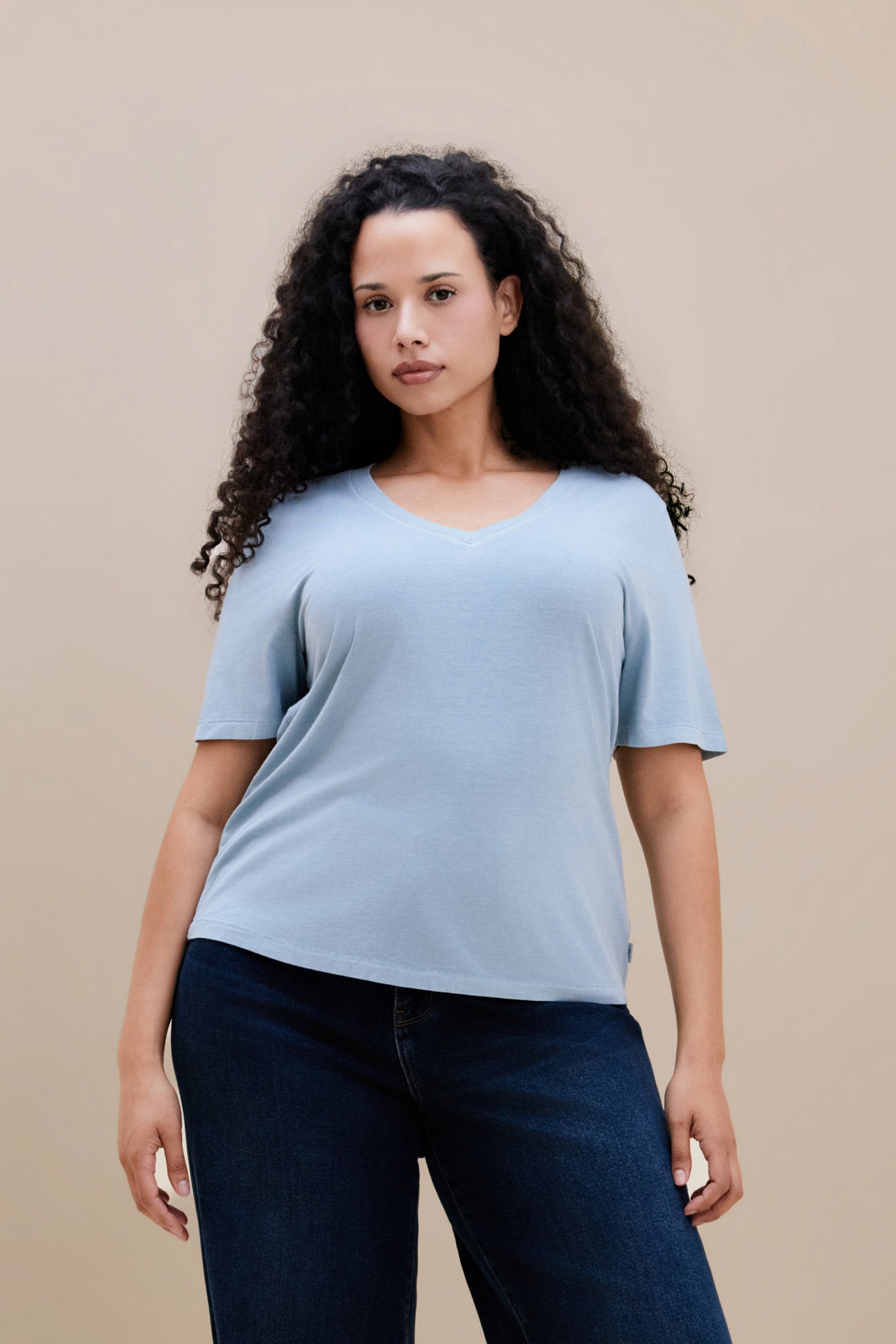 Woman wearing a light blue t-shirt and dark jeans against a beige background | model-size: 16
