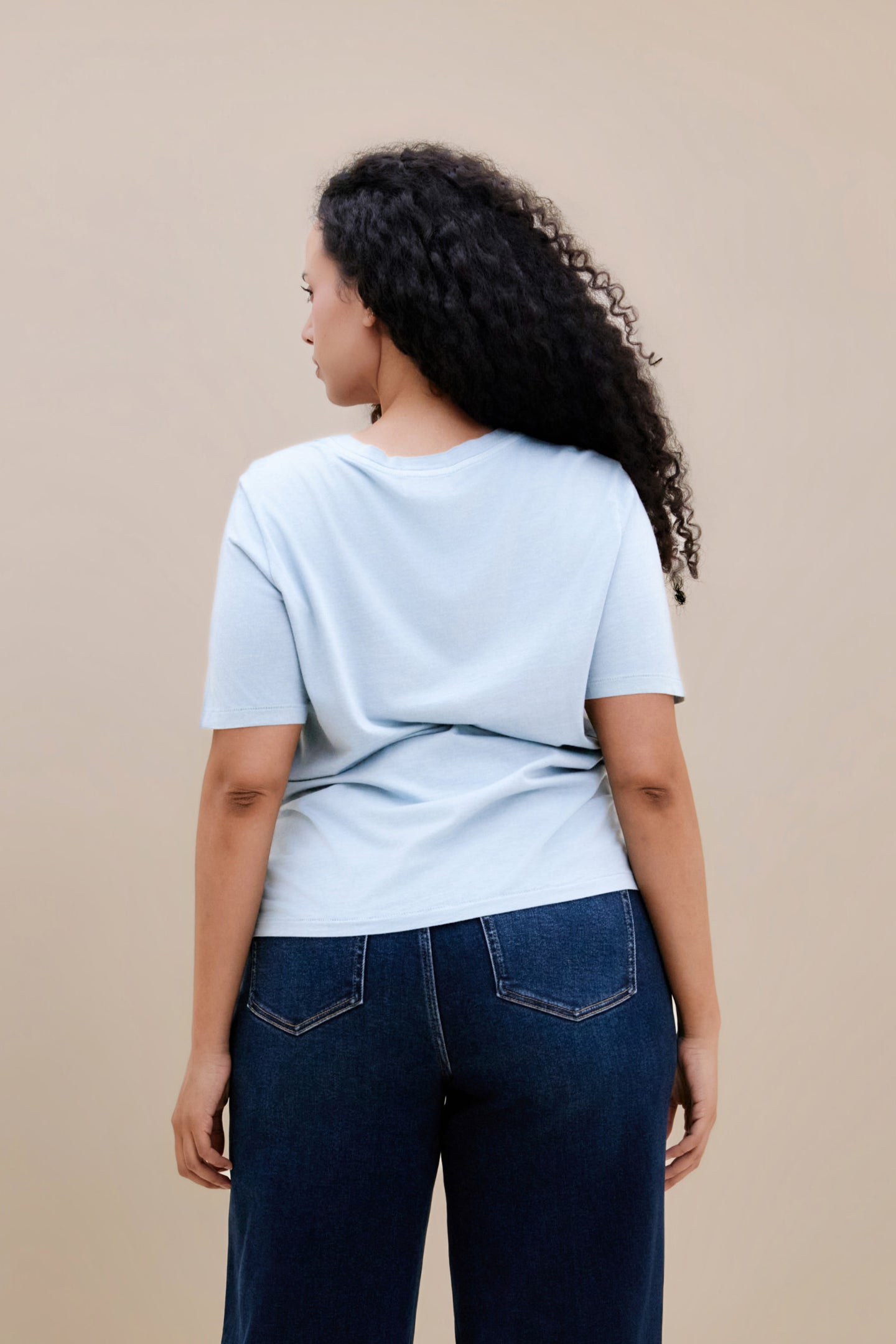 Backside of a woman wearing a light blue t-shirt and dark jeans against a beige background