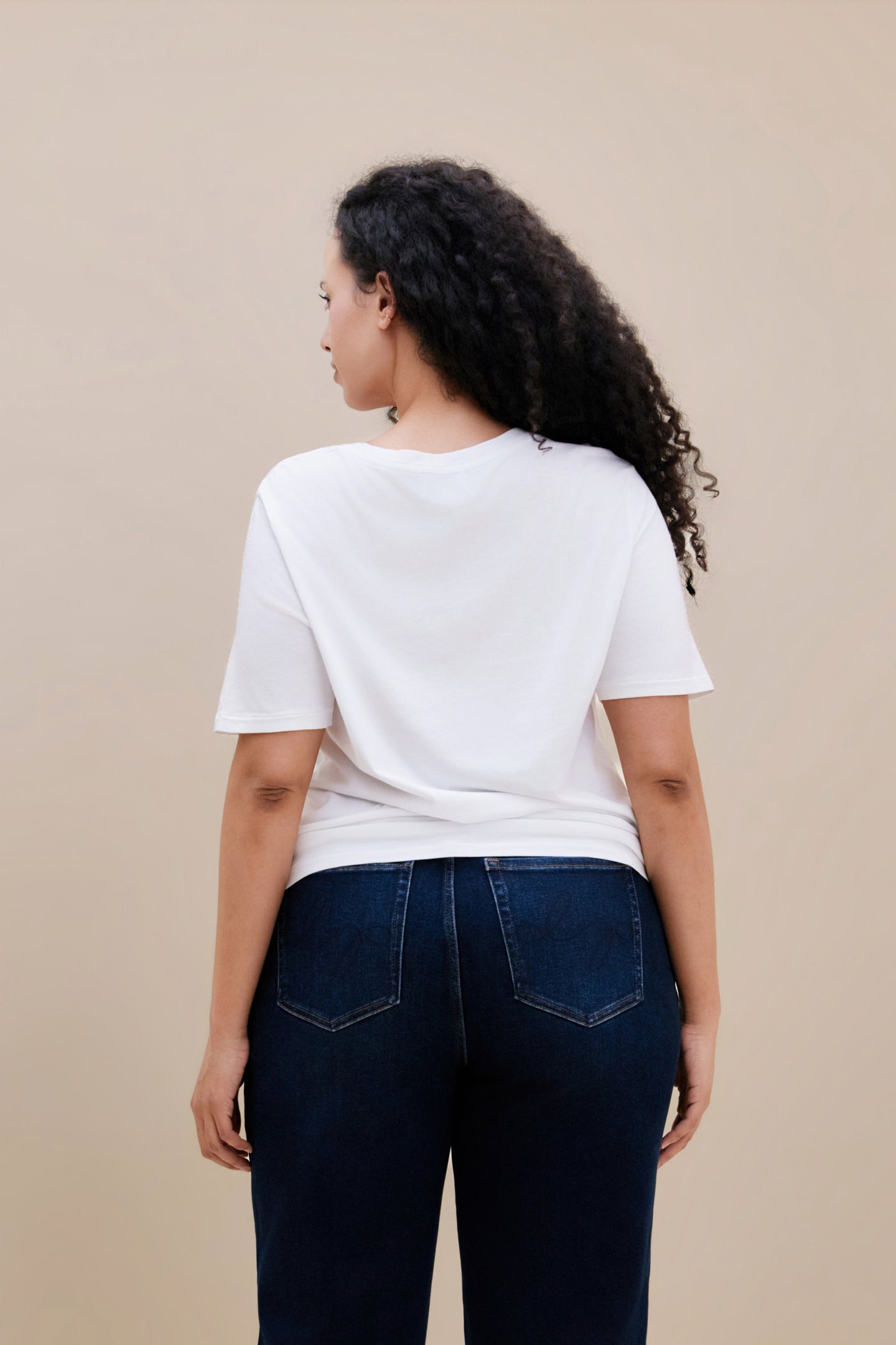 Woman wearing a plain white t-shirt and dark jeans against a beige background