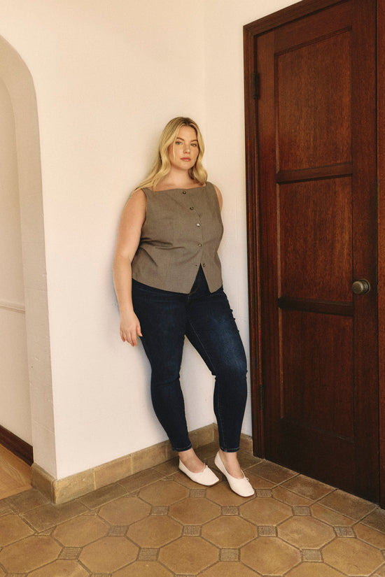 ADA Complaint: Shop Skinny Jeans.  Woman standing inside leaning against a white wall wearing sleeveless gray button up to and dark blue jeans.