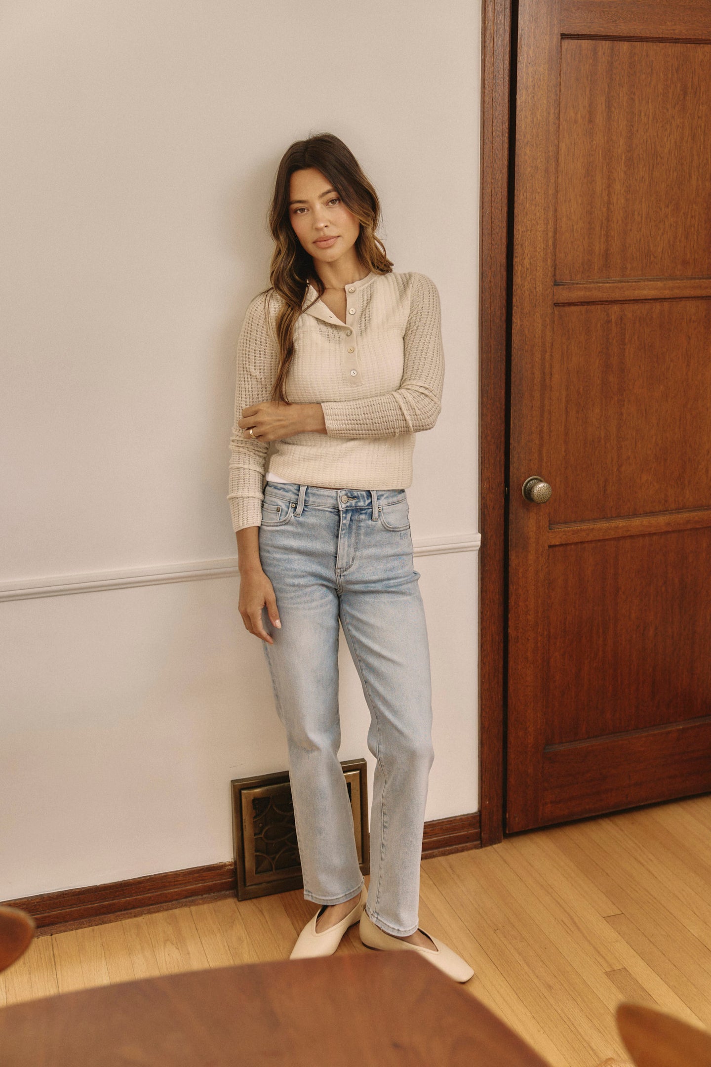 Woman standing indoors wearing a cream sweater and light blue jeans.