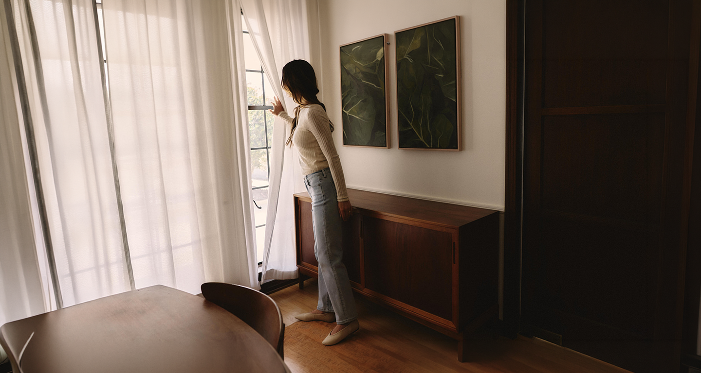 Woman standing in a dining room looking out a window wearing light jeans and a white top