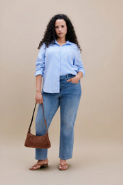Woman wearing a blue button down and jeans holding a purse with a beige background | model-size: 16