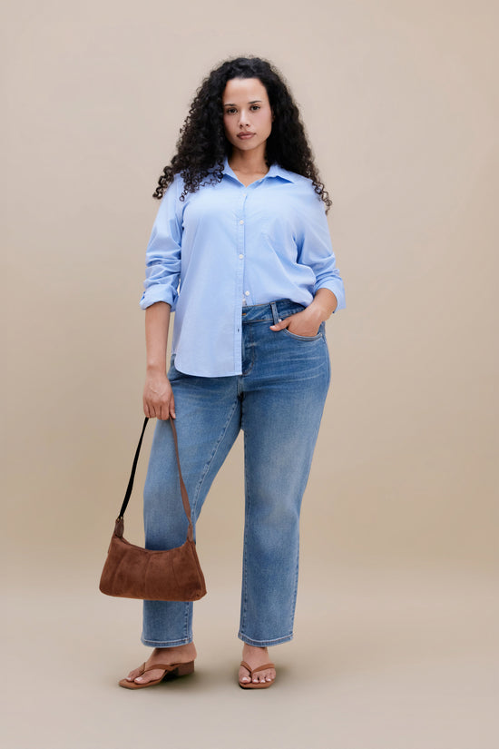 Woman wearing a blue button down and jeans holding a purse with a beige background | model-size: 16