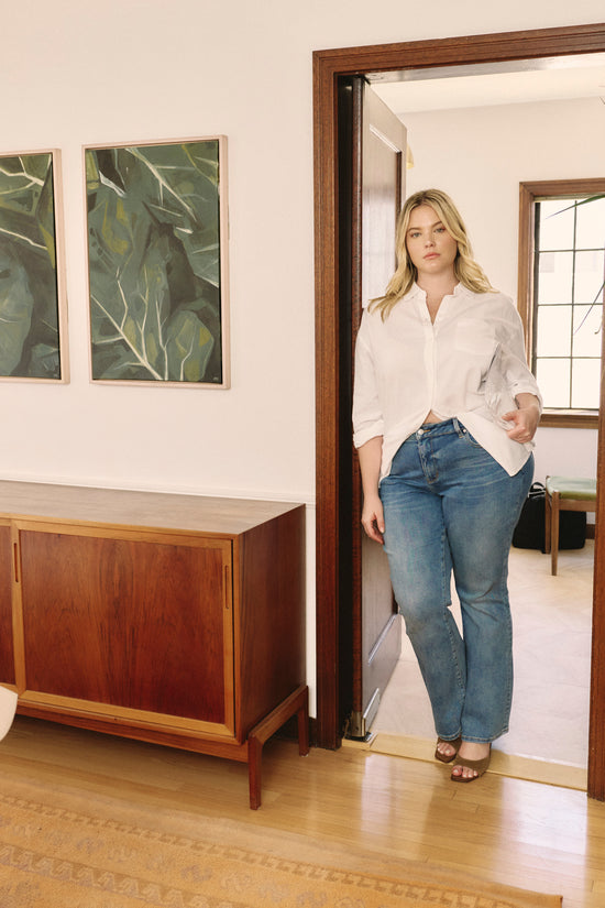 Woman standing in a room with wooden floor and wall art wearing a white button up blouse and jeans.