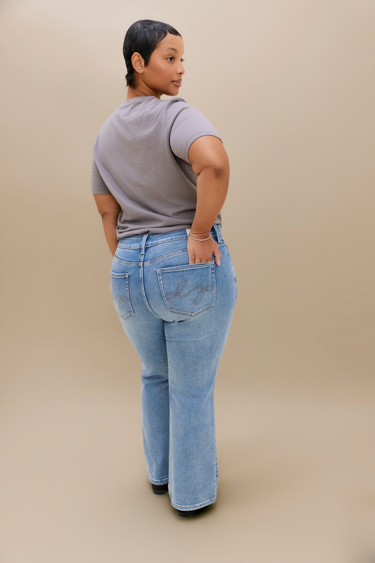 A woman shown from the back wearing light-wash blue boot cut jeans and a gray t-shirt, standing against a solid beige studio background.