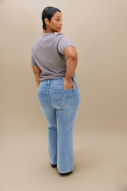 A woman shown from the back wearing light-wash blue boot cut jeans and a gray t-shirt, standing against a solid beige studio background.