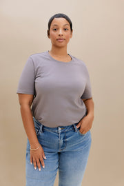 A woman modeling a short-sleeve crewneck t-shirt in a muted grayish-purple color, paired with blue jeans, against a solid beige background.