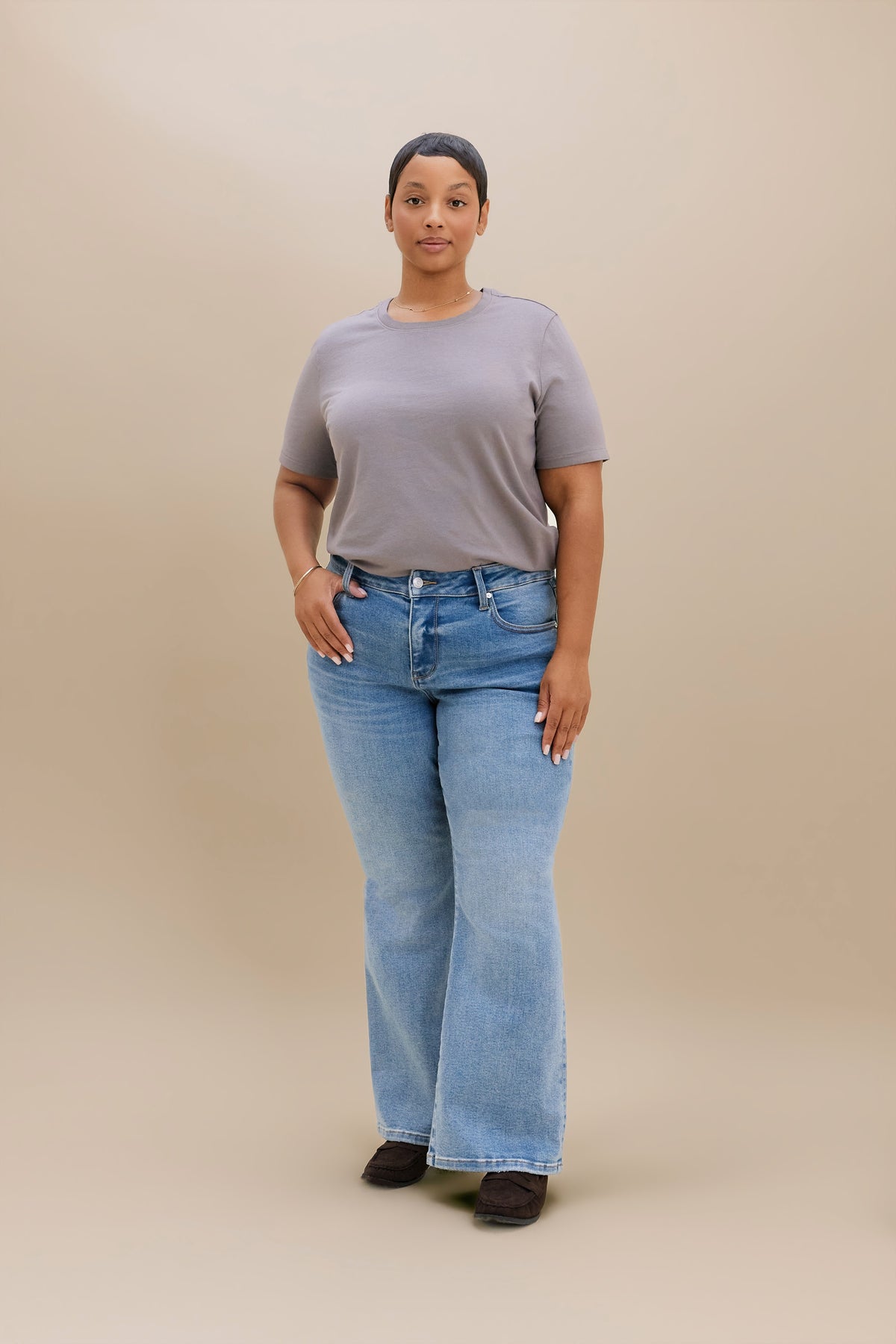 A woman stands against a plain beige studio background wearing a neutral colored t-shirt light blue boot cut jeans, dark brown heeled boots.