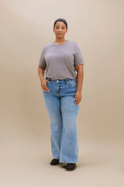 A woman stands against a plain beige studio background wearing a neutral colored t-shirt light blue boot cut jeans, dark brown heeled boots.