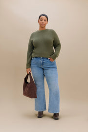 A model stands against a solid beige studio background wearing an olive green long-sleeve knit sweater, light-wash blue boot cut jeans, and dark brown boots, while holding a dark brown suede tote bag.