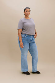 A woman modeling light-wash blue flare jeans with a taupe-gray t-shirt, standing against a solid beige studio background.