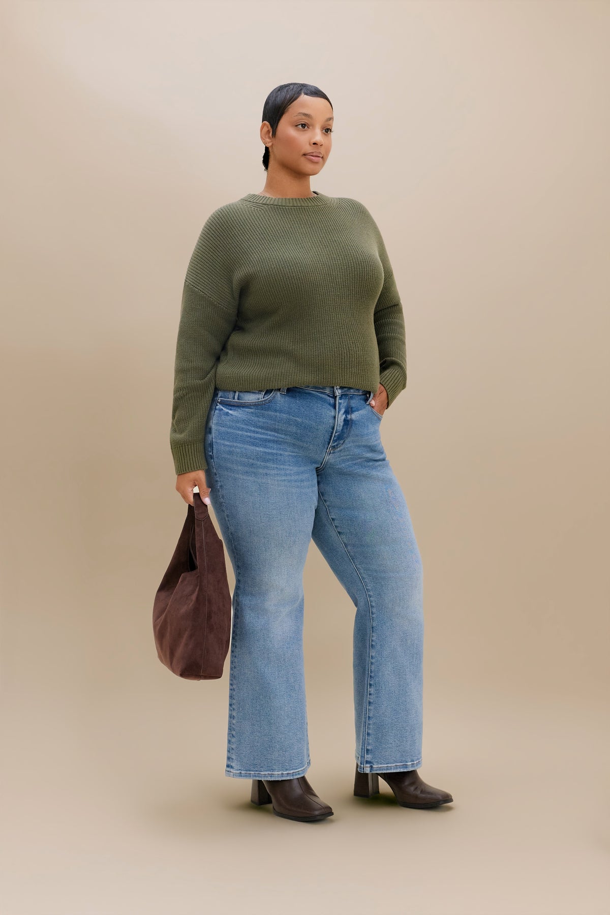 A woman stands against a plain beige studio background wearing an olive green long-sleeve knit sweater, light blue boot cut jeans, dark brown heeled boots, and holding a brown tote bag.