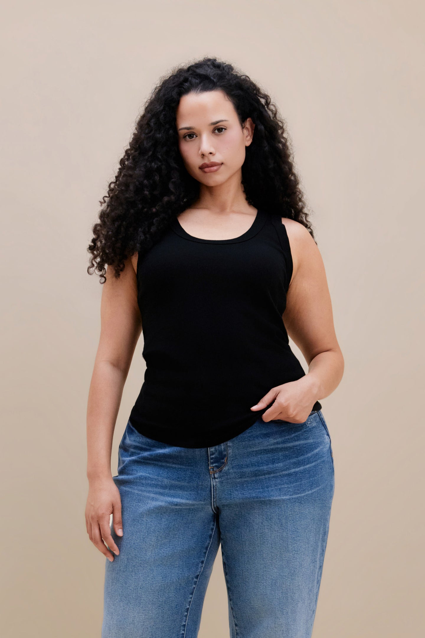 Woman wearing a black ribbed tank top and jeans against a beige background | model-size: 16