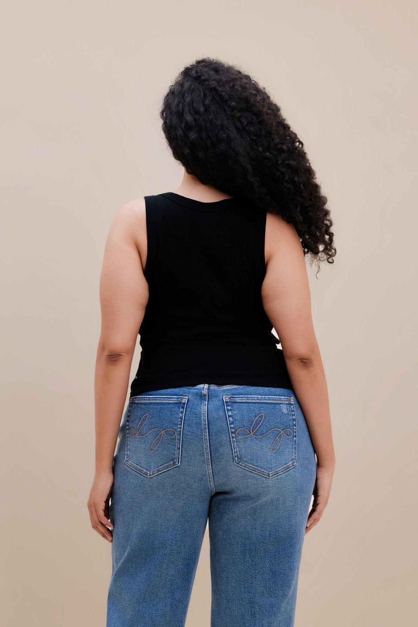 Backside of a woman wearing a black ribbed tank top and jeans against a beige background