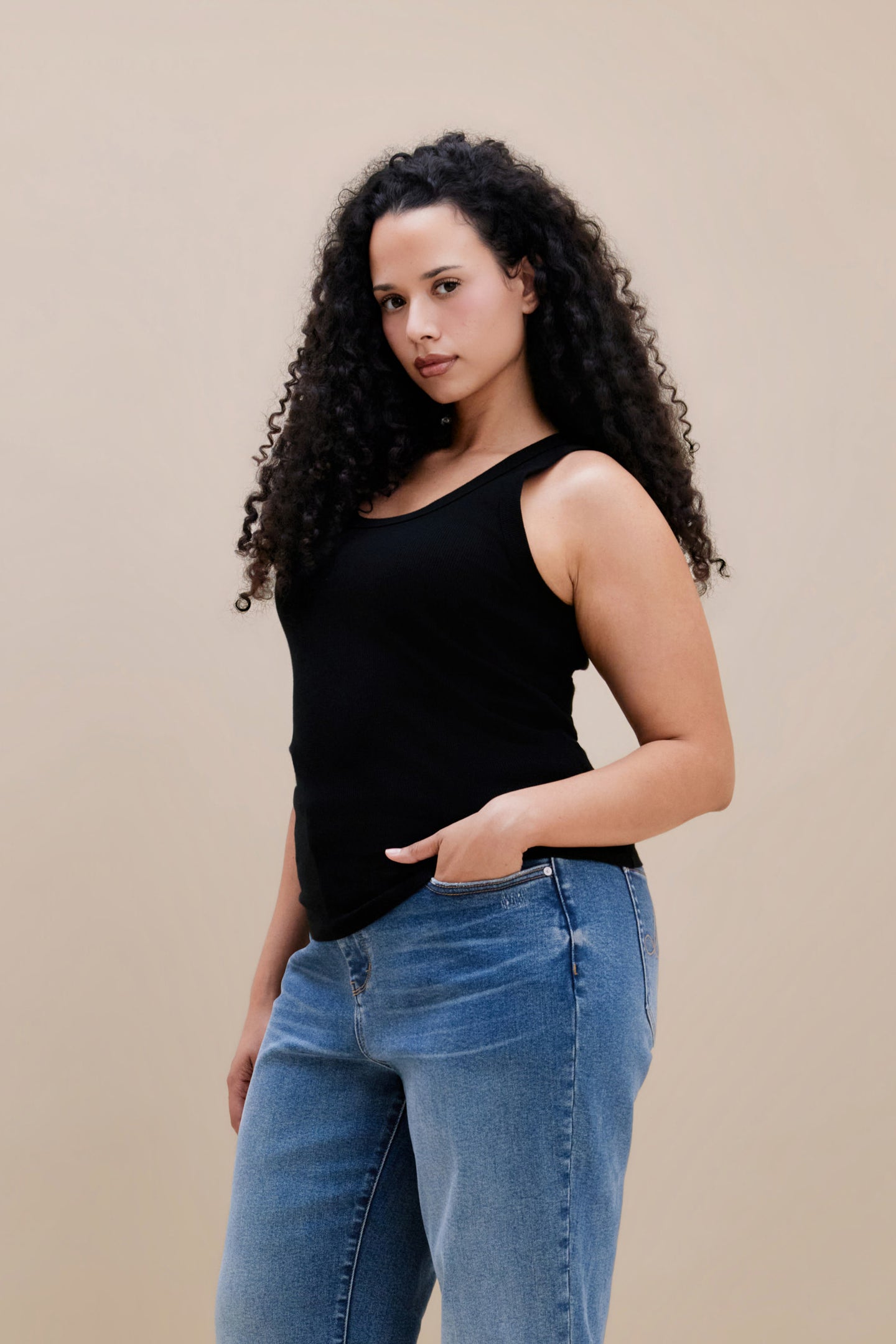 Woman wearing a black ribbed tank top and jeans against a beige background
