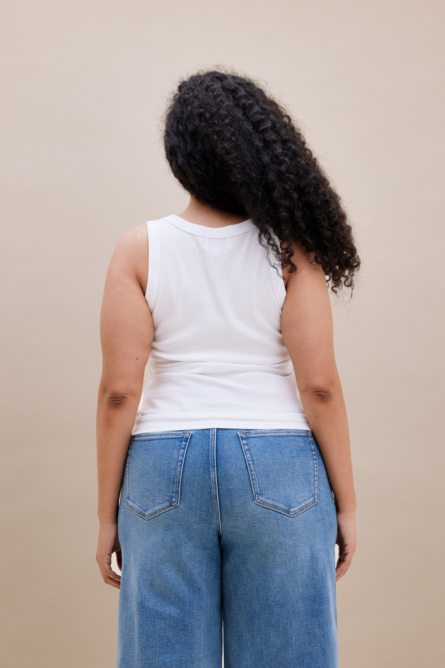 Backside of a woman wearing a white ribbed tank top and jeans against a beige background