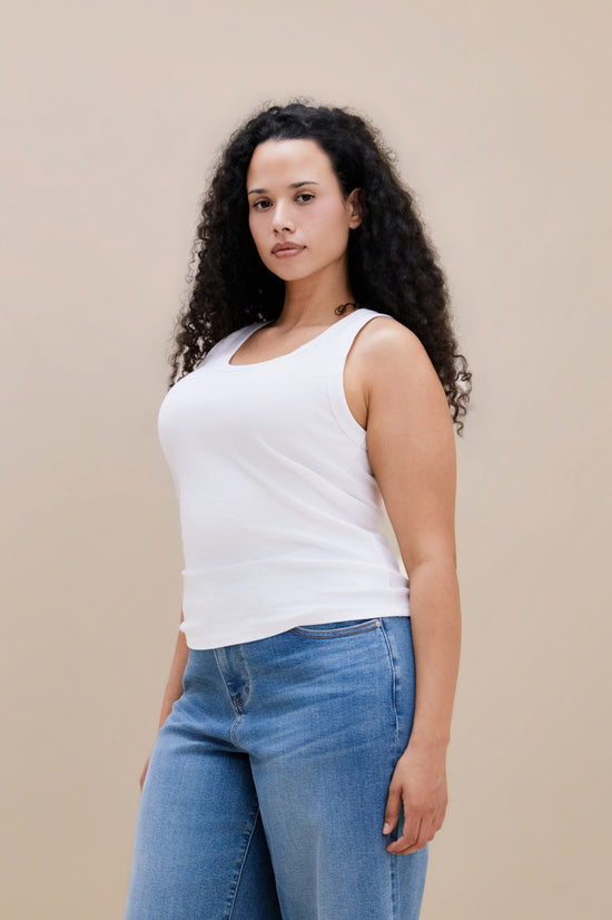 Woman wearing a white ribbed tank top and jeans against a beige background