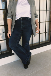 Person wearing dark blue jeans and a gray coat standing against a windowed wall | model-size: 16