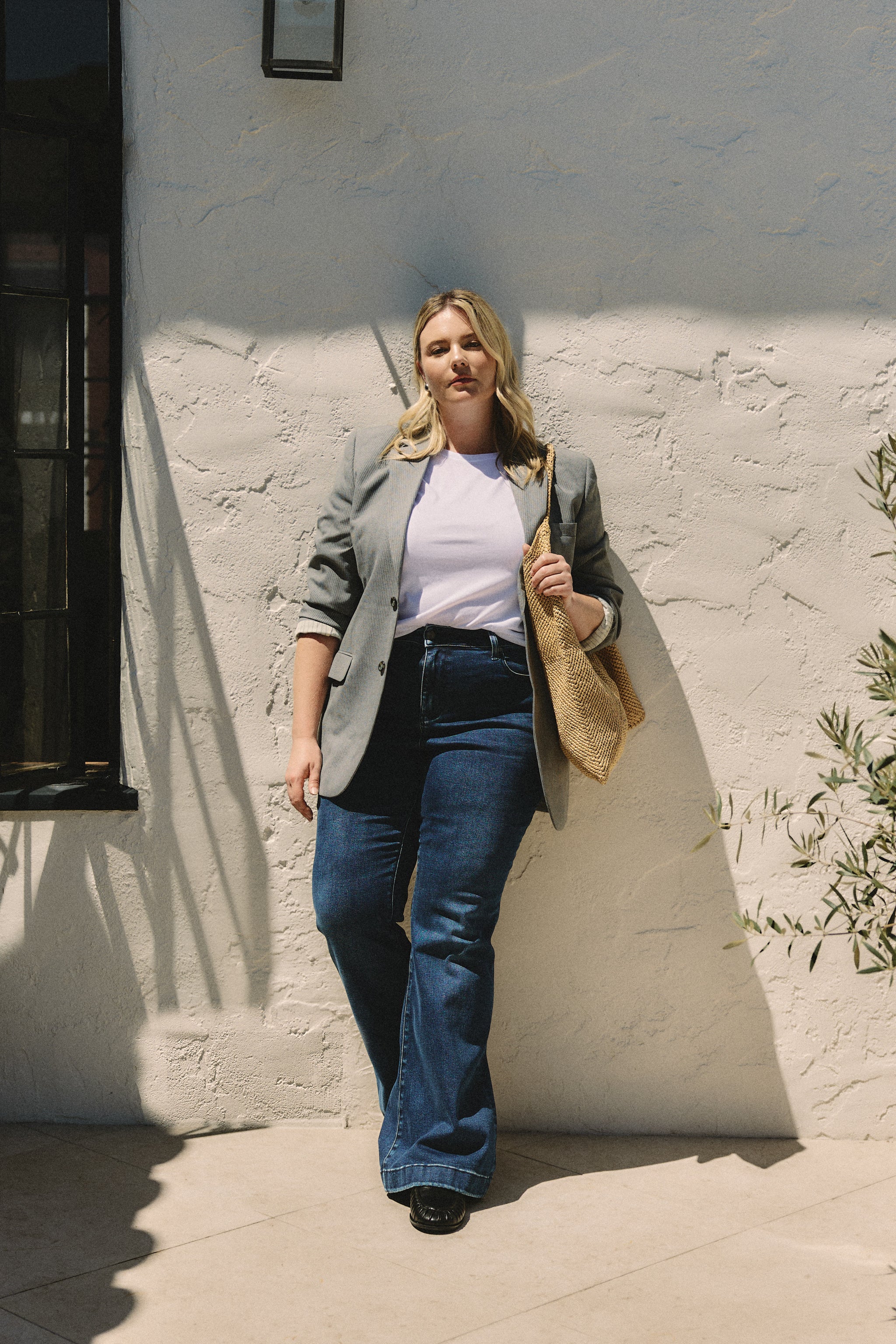 Woman standing against a white wall wearing a gray blazer, white shirt, and blue jeans.