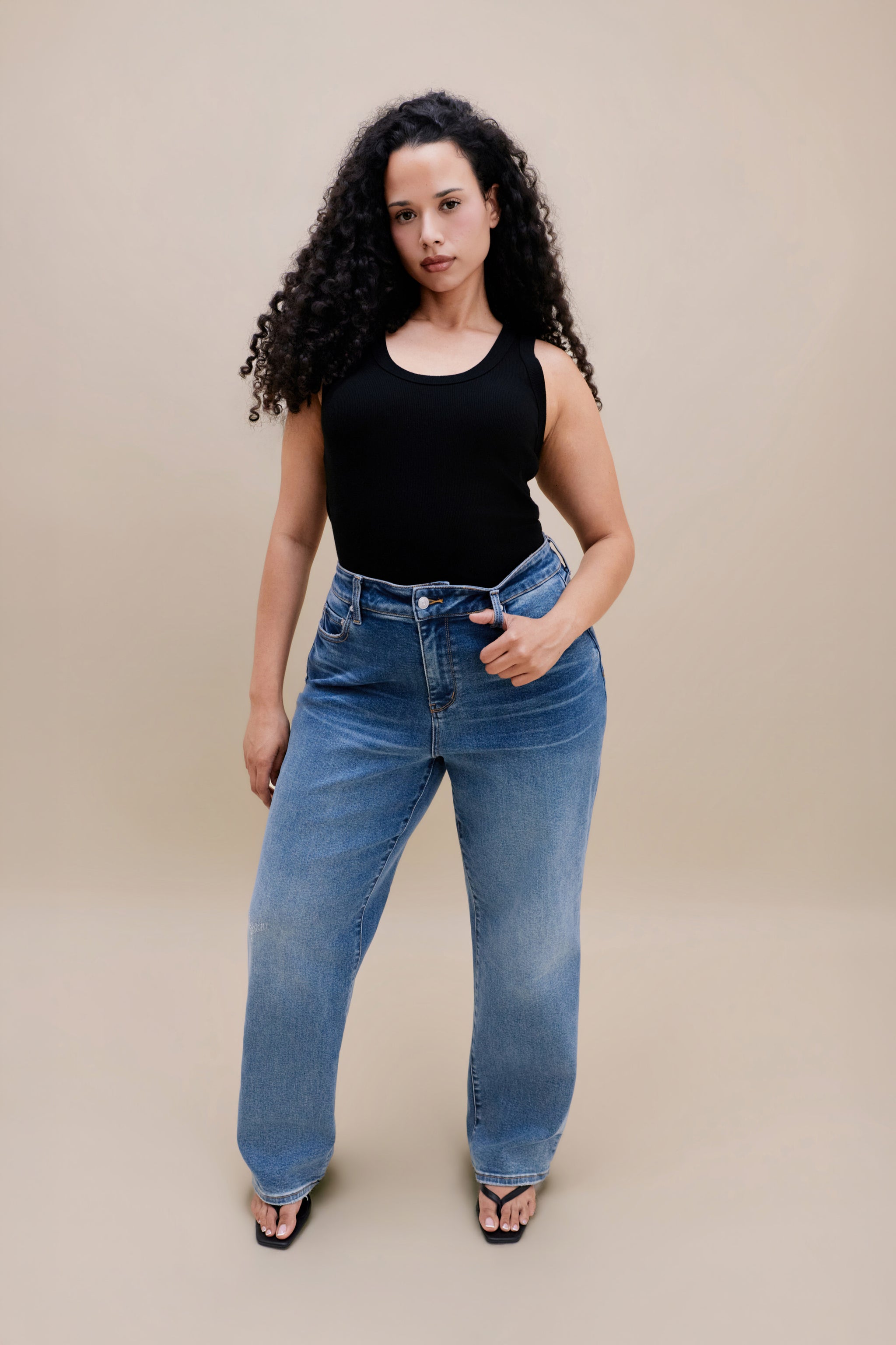 Woman wearing a black tank top and jeans with a beige background | model-size: 16
