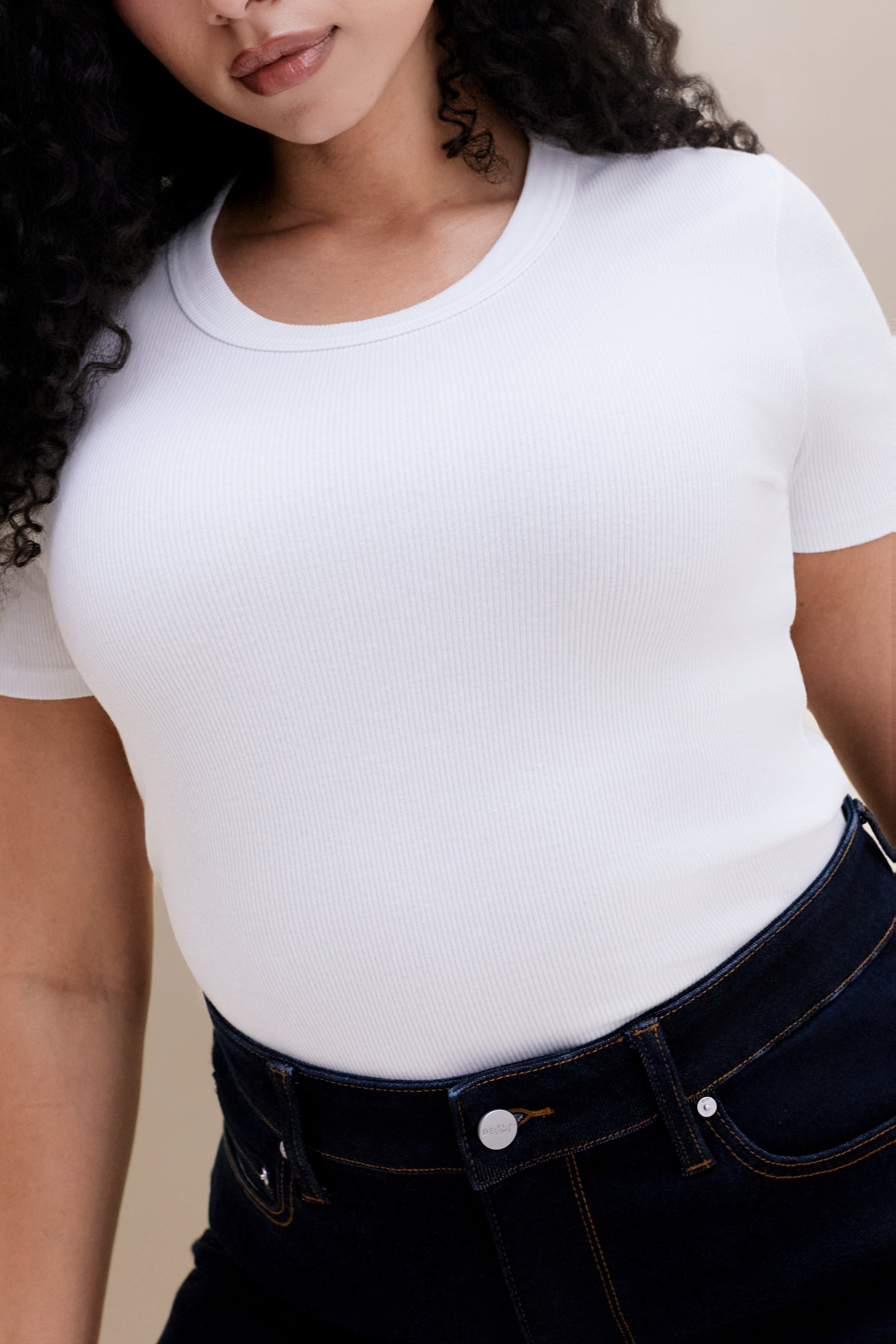 Person wearing a plain white t-shirt and dark jeans on a beige background