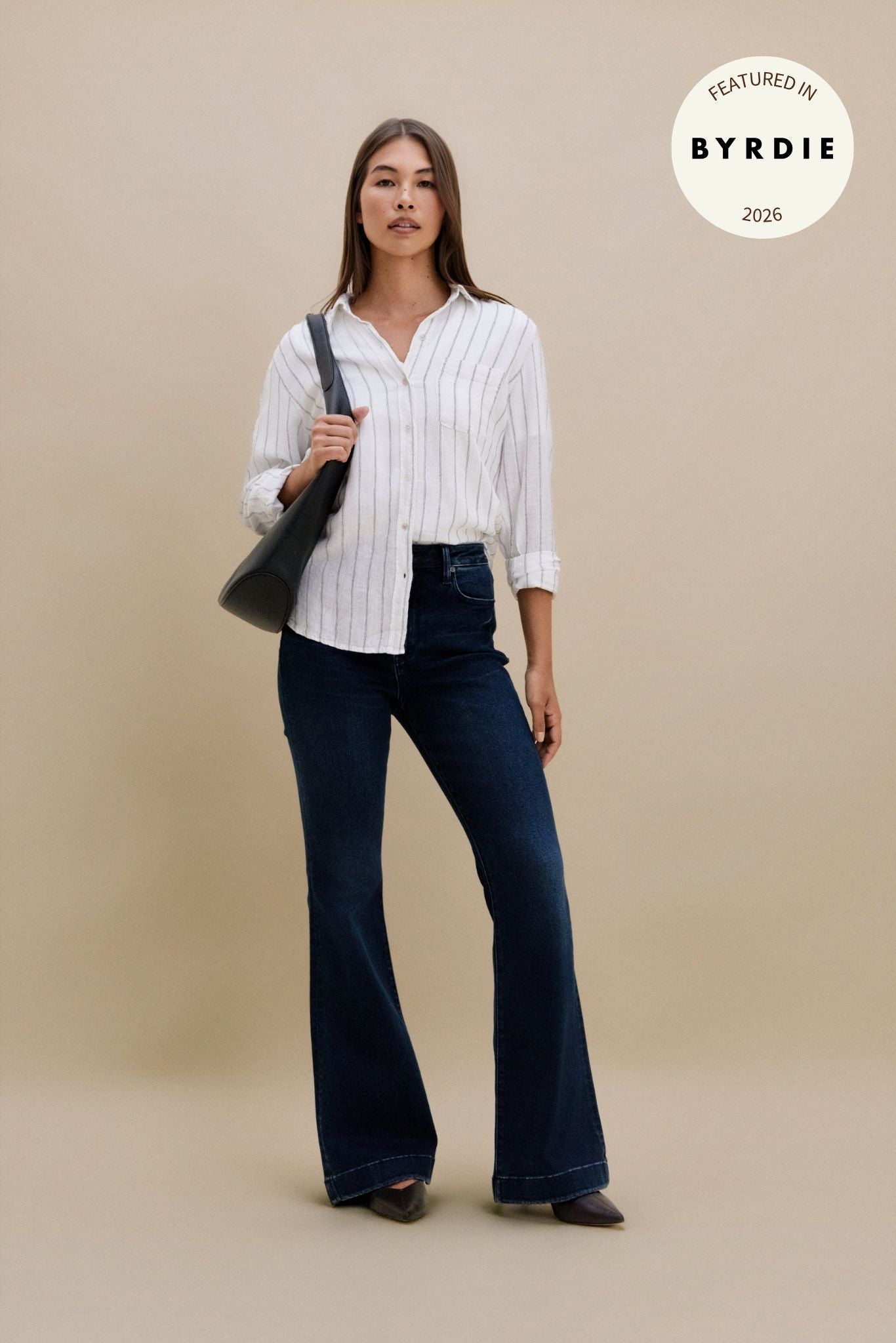 Woman wearing a white striped shirt and dark jeans, holding a black bag on a beige background with 'Byrdie' logo | model-size: 4