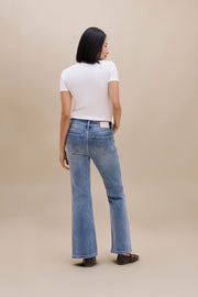 A woman is shown from behind wearing light-wash blue flare jeans, a white short-sleeve t-shirt, and dark brown flat shoes against a solid beige studio background.
