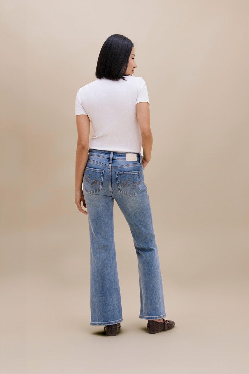 A woman is shown from behind wearing light-wash blue flare jeans, a white short-sleeve t-shirt, and dark brown flat shoes against a solid beige studio background.