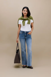 A woman stands against a plain beige studio background wearing a white short-sleeve top, blue high-waisted flare jeans, and dark flats. She has an olive green knit wrap tied around her shoulders and is holding a dark taupe tote bag.