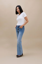 A woman modeling a pair of light-wash blue flare jeans with a white short-sleeve top, standing against a solid beige studio background.