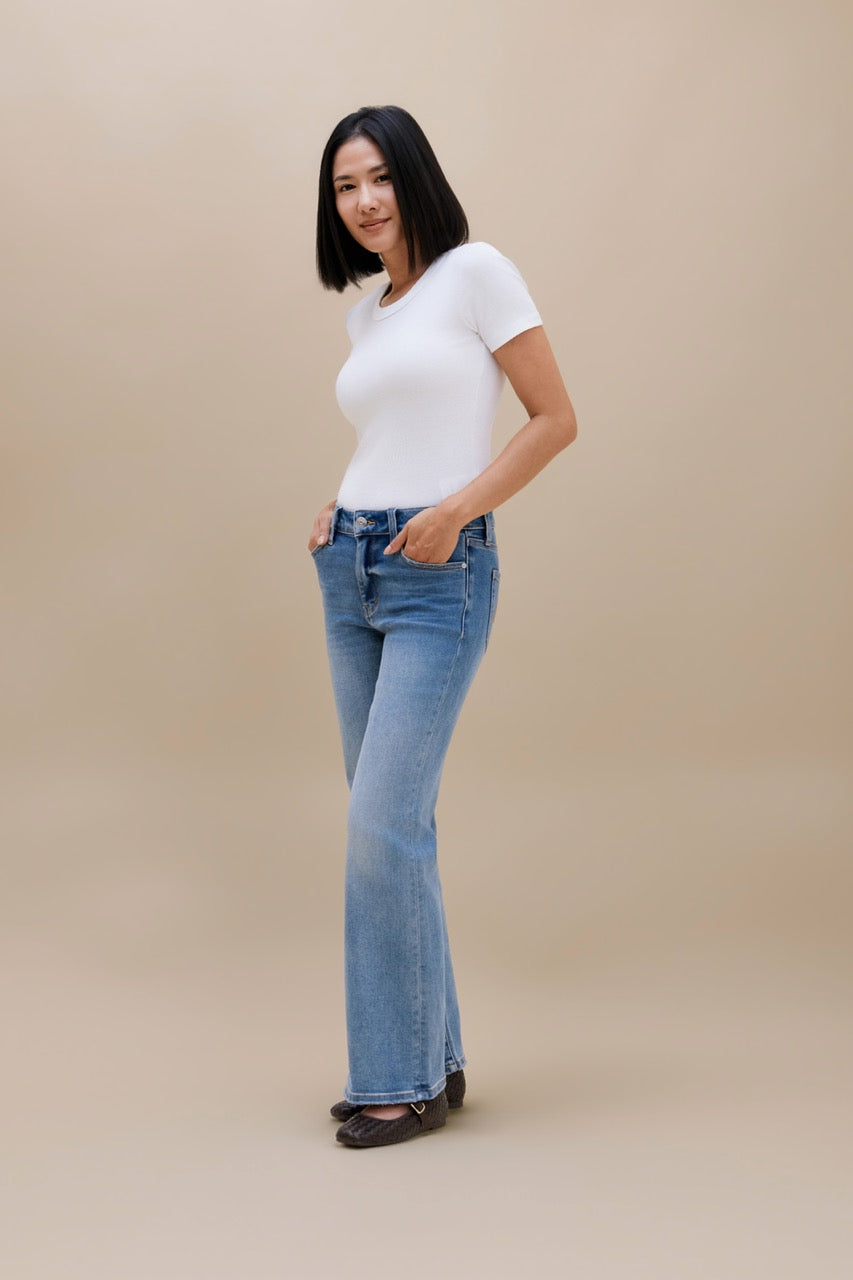 A woman modeling a pair of light-wash blue flare jeans with a white short-sleeve top, standing against a solid beige studio background.