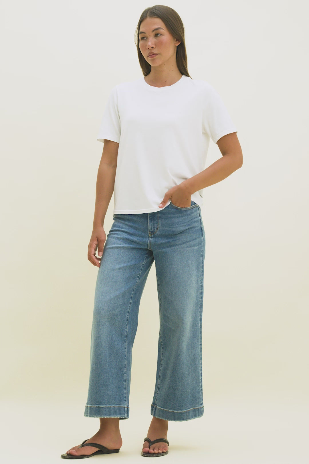 Video of a woman wearing a white t-shirt and ankle length wide leg blue jeans with a beige background.