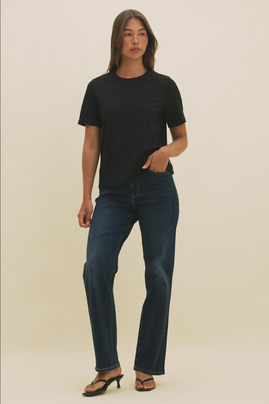 Video of a woman wearing a black t-shirt and dark straight leg jeans with a beige background.