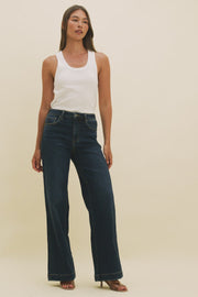 Video of a woman wearing a white tank top and dark blue wide leg jeans.